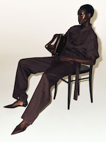 Placeholder for: A person in a dark brown outfit sits on a chair, holding a brown bag, with legs casually stretched out and wearing pointed shoes.