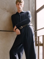 Placeholder for: Person in a dark, loose-fitting outfit leans against a wall by a window, with a relaxed pose and neutral expression.