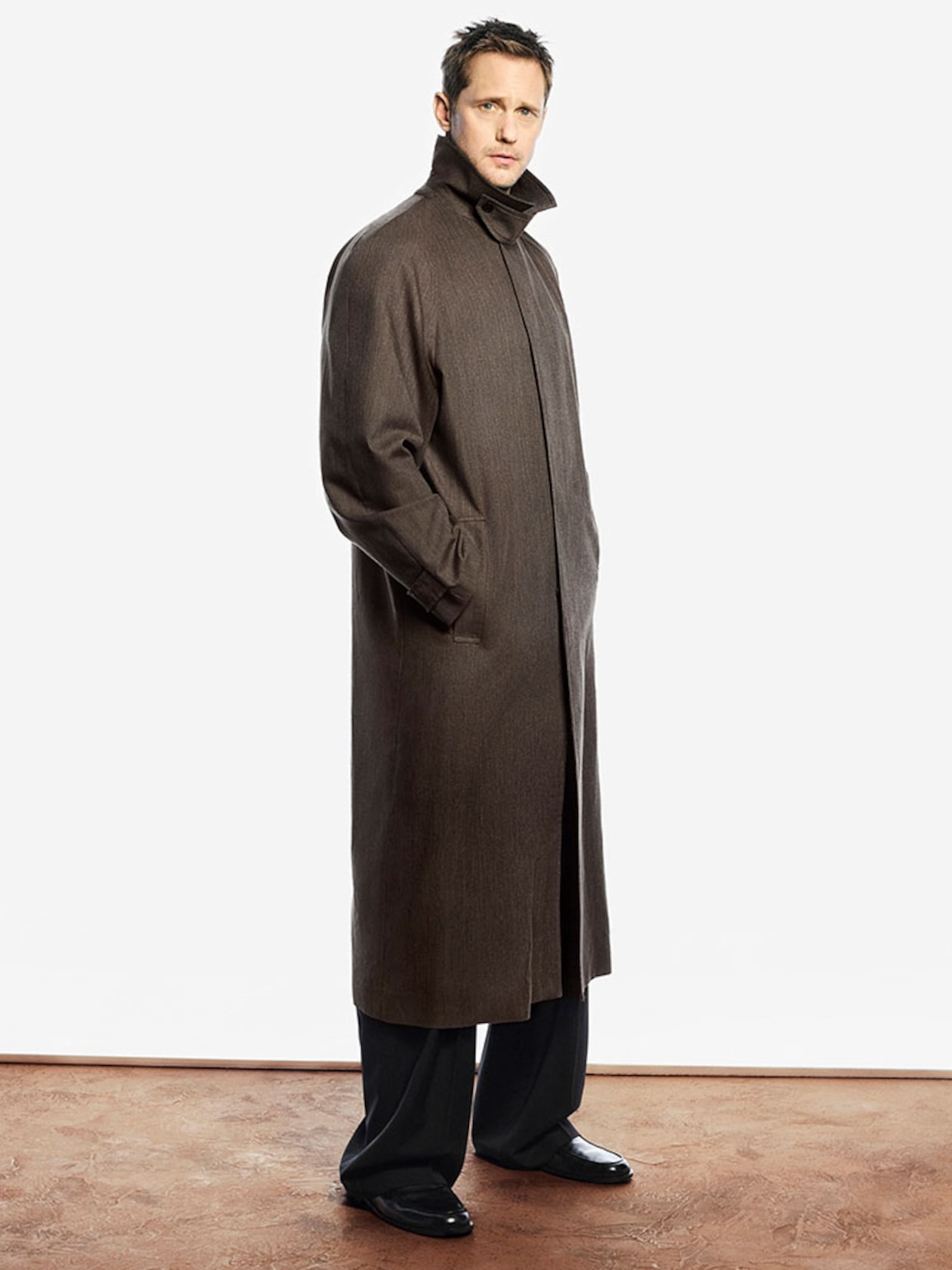 Person wearing a long brown coat with a high collar, dark pants, and black shoes, standing against a plain white background.