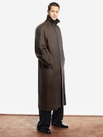 Placeholder for: Person wearing a long brown coat with a high collar, dark pants, and black shoes, standing against a plain white background.