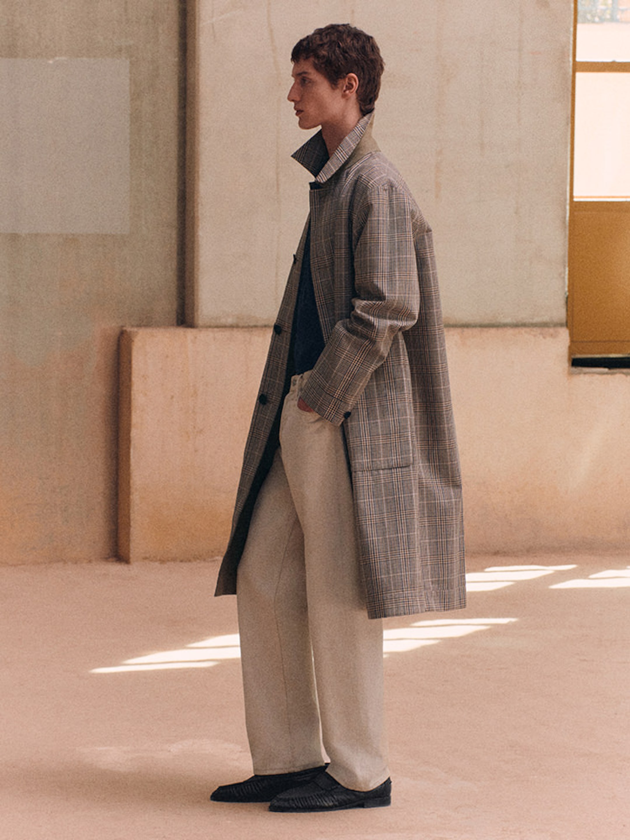 A person in profile wears a long, checked coat over light pants, standing in a minimalist room with soft, neutral tones.