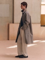 Placeholder for: A person in profile wears a long, checked coat over light pants, standing in a minimalist room with soft, neutral tones.
