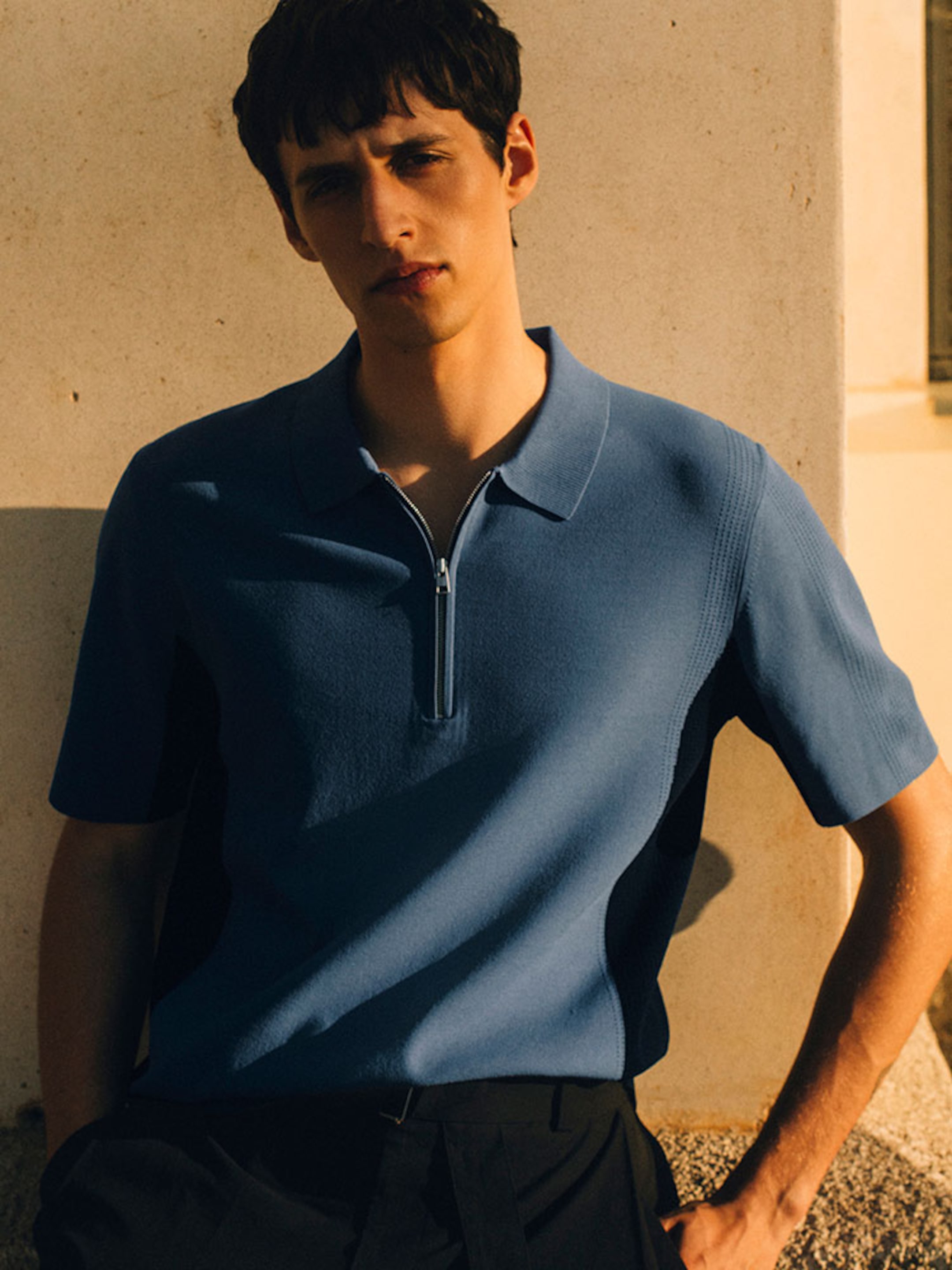 A medium-shot portrait of a man standing against a warm, sunlit stone wall during the golden hour. He is wearing a blue short-sleeved knit polo shirt featuring a silver-tone quarter-zip neckline and contrasting dark side panels. The shirt is styled with pleated black tailored trousers, emphasizing a refined yet relaxed summer holiday aesthetic.