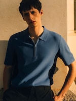 Placeholder for: A medium-shot portrait of a man standing against a warm, sunlit stone wall during the golden hour. He is wearing a blue short-sleeved knit polo shirt featuring a silver-tone quarter-zip neckline and contrasting dark side panels. The shirt is styled with pleated black tailored trousers, emphasizing a refined yet relaxed summer holiday aesthetic.