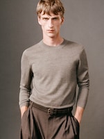Placeholder for: A person with short hair wearing a gray sweater and dark pants stands against a neutral background, looking directly at the camera.