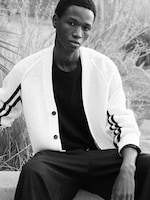 Placeholder for: Person seated outside wearing a white cardigan with black stripes over a black shirt, looking at the camera, with plants in the background.