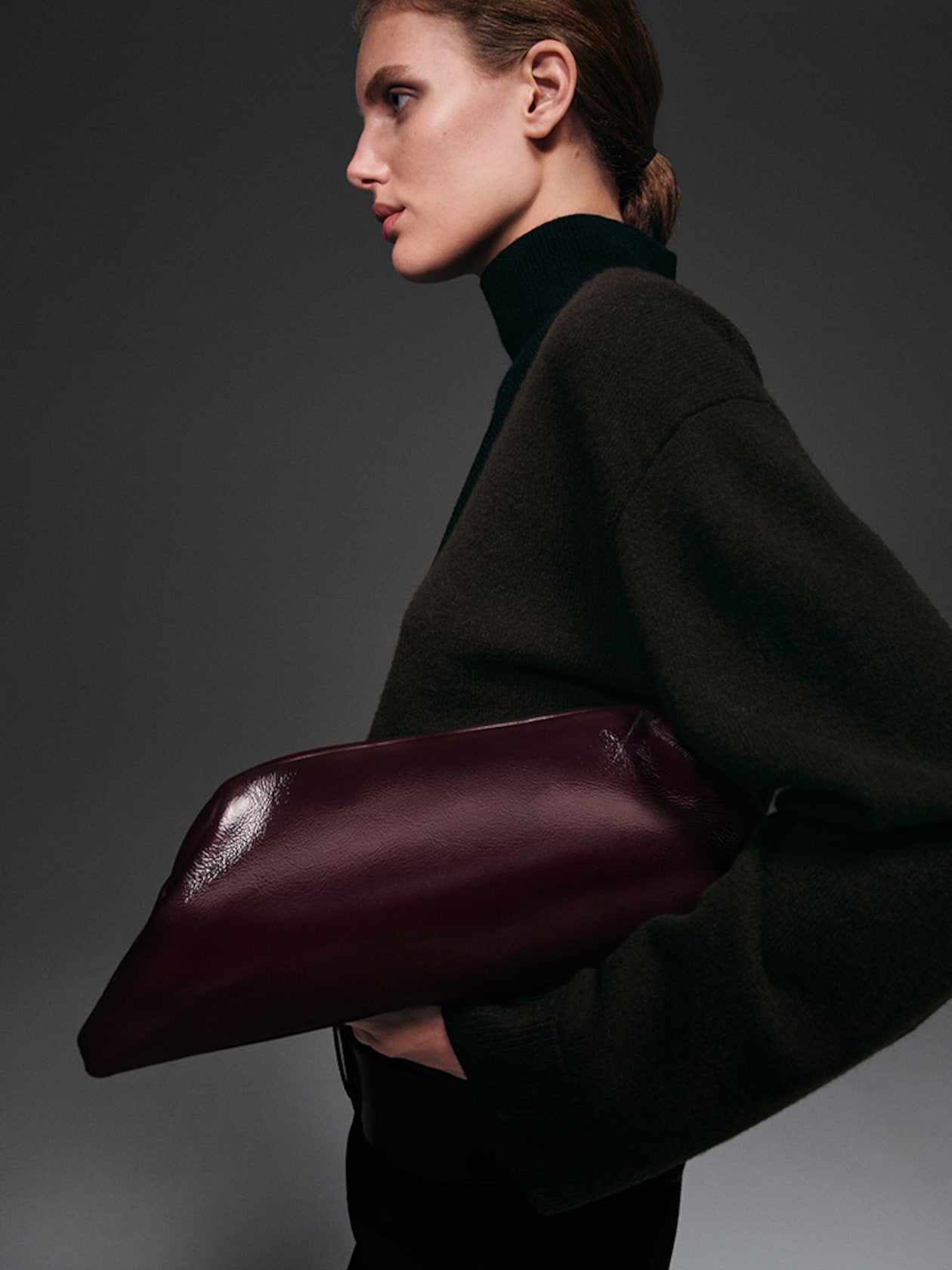 Profile of a person in a dark turtleneck holding a large, glossy burgundy clutch against a dark background.