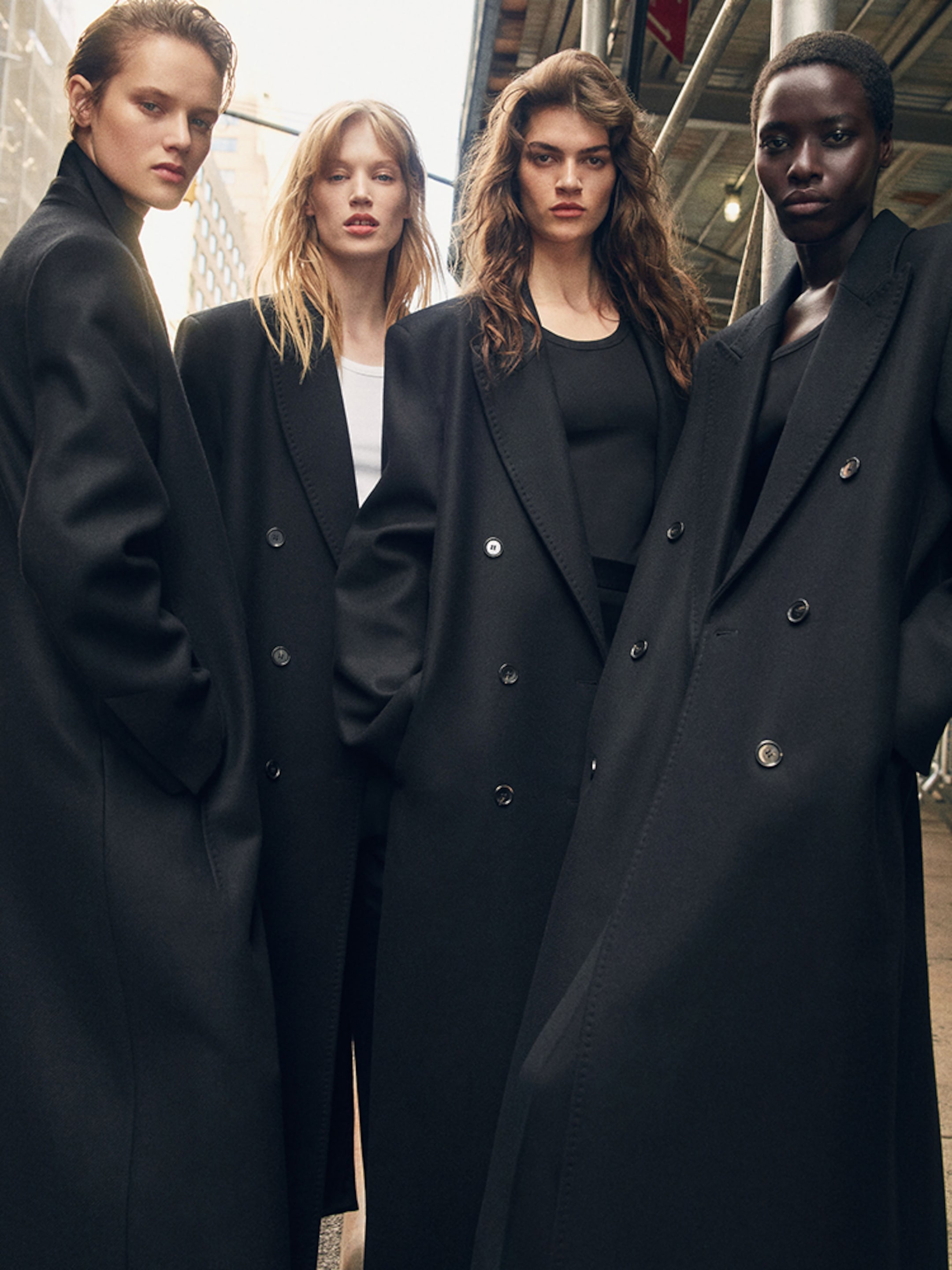 Four models in black long coats stand confidently in an urban setting, framed beneath a scaffolding with city buildings in the background.