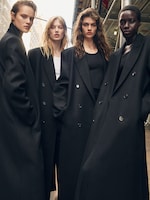 Placeholder for: Four models in black long coats stand confidently in an urban setting, framed beneath a scaffolding with city buildings in the background.