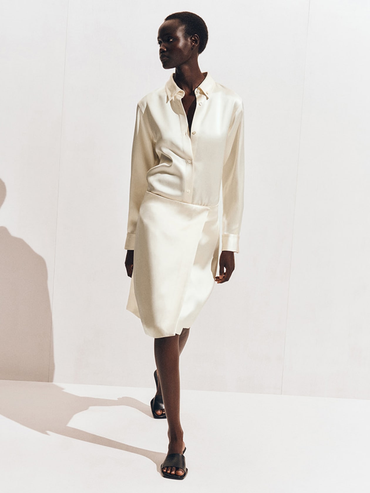 A woman wearing a cream-colored, long-sleeve silk shirt and skirt, walking confidently on a white background with shadows.