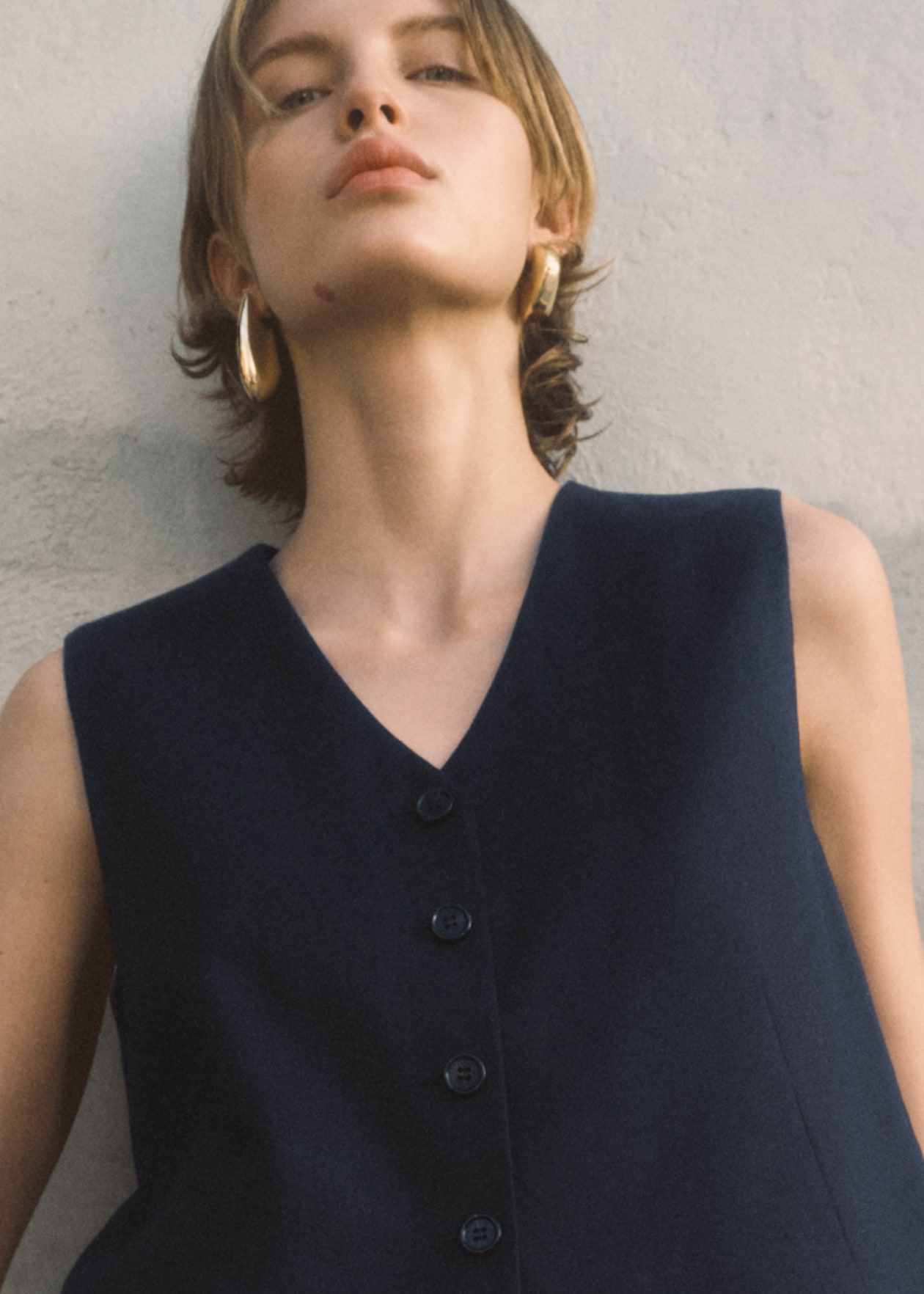 Model leaning against the wall. She's wearing golden hoop earrings and is wearing a black top.