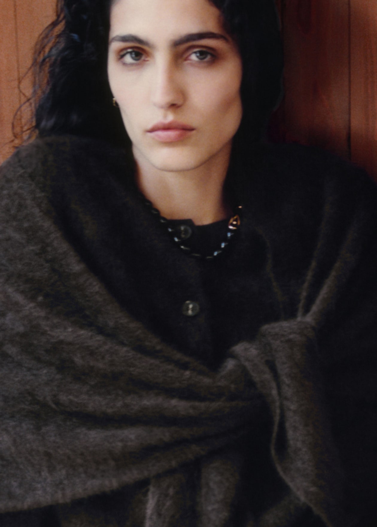 Person with long dark hair wearing a dark, textured shawl and a necklace, looking at the camera against a wooden background.