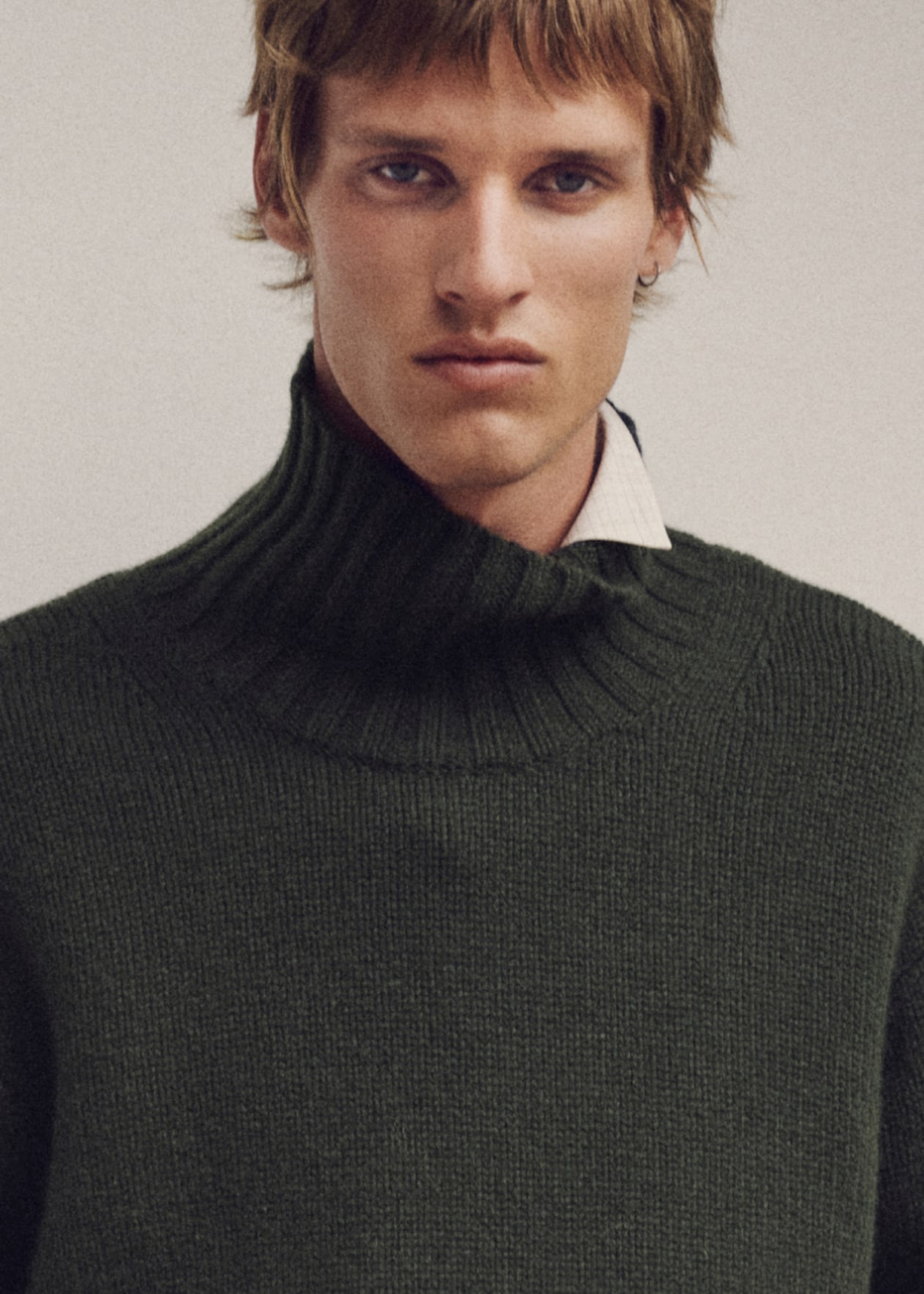 Person wearing a dark green turtleneck sweater over a white shirt, with short hair, looking directly at the camera against a plain background.