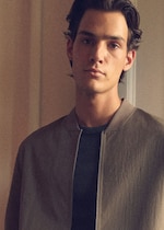 Placeholder for: Model with dark hair wearing a light brown jacket and a dark shirt stands in softly lit indoor surroundings, looking at the camera.