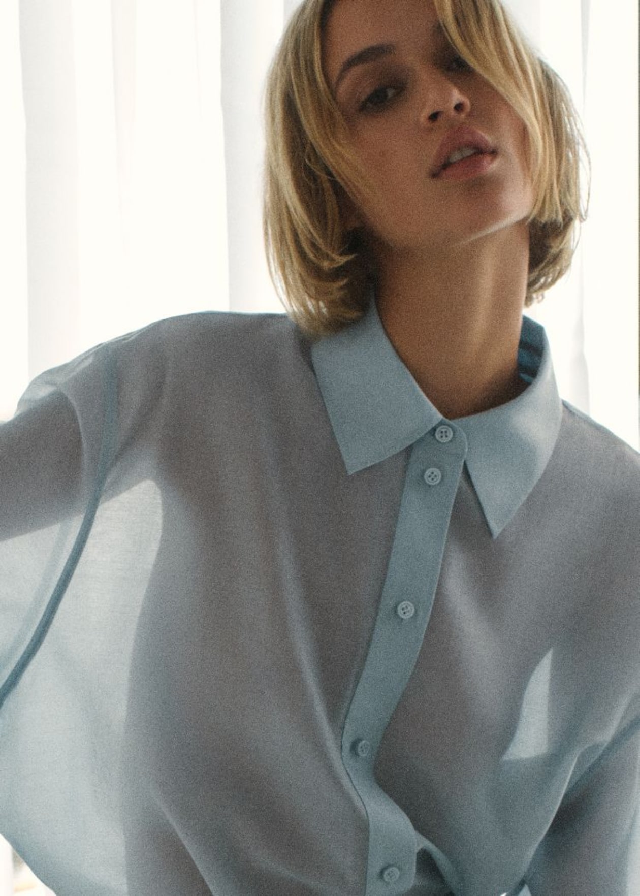 Model with short blond hair wearing a sheer blue button-up shirt stands in front of softly lit white curtains.