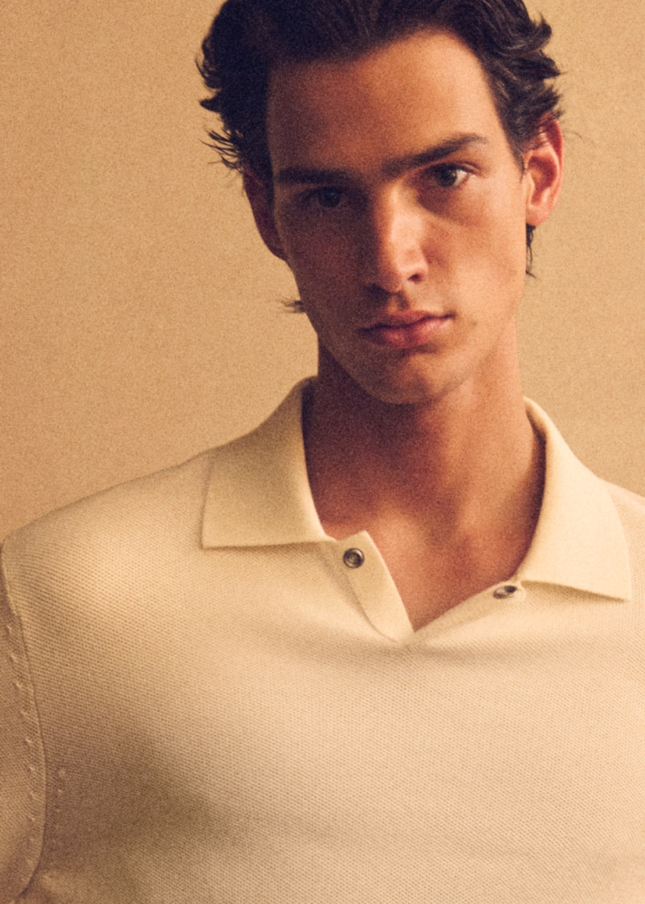 A person wearing a cream-colored polo shirt, looking directly at the camera with a neutral expression and a beige background.