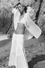 Placeholder for: Woman in a white outfit poses on a rocky beach, wearing a shirt and skirt, with arms raised, eyes closed, and waves in the background. Black and white photo.