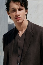 Placeholder for: Young man with dark hair wearing a brown suit, standing outdoors against a light background, looking into the distance.
