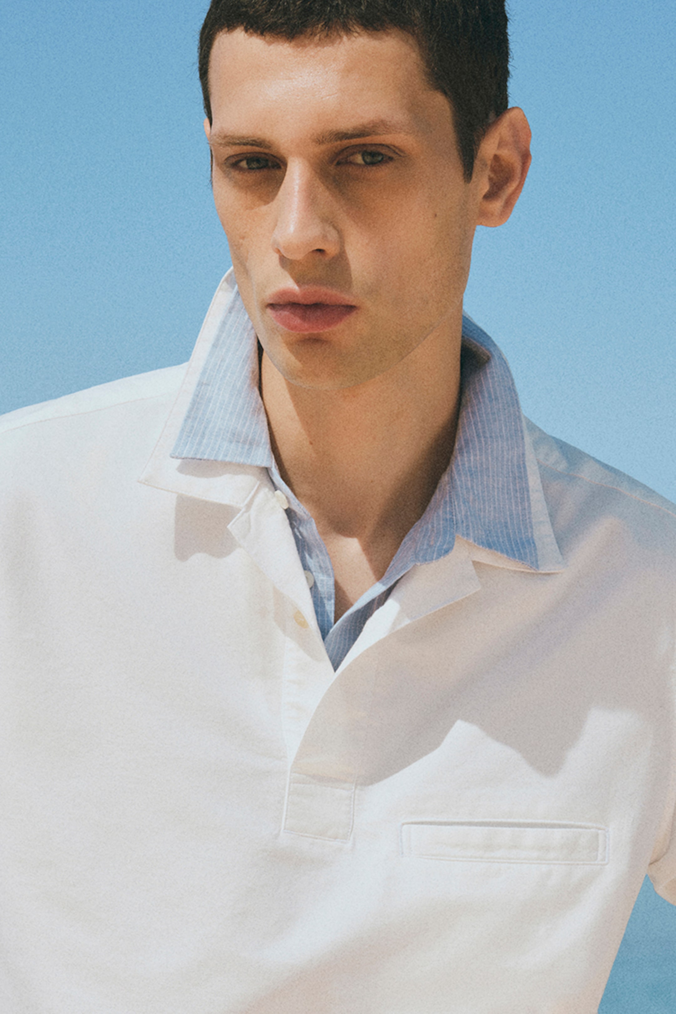 Man in a white shirt with a blue collar stands against a clear blue sky, looking serious.