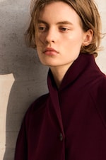 Placeholder for: Person with short hair wearing a dark maroon coat, standing against a textured wall, gazing thoughtfully to the side.