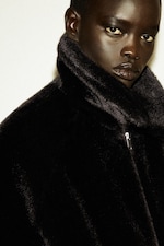 Placeholder for: Person wearing a dark, high-collared fur coat, gazing intently at the camera with a neutral expression.