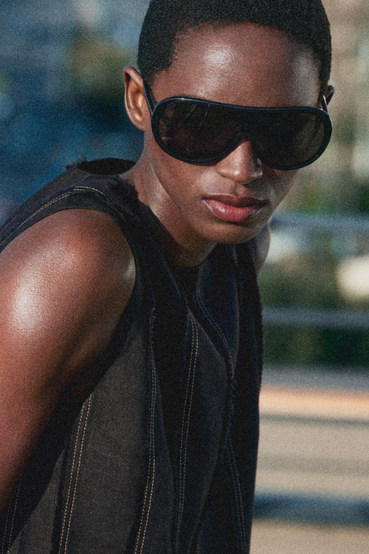 Person wearing large black sunglasses and a dark sleeveless top, looking confidently towards the camera, with a blurred outdoor background.