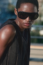 Placeholder for: Person wearing large black sunglasses and a dark sleeveless top, looking confidently towards the camera, with a blurred outdoor background.