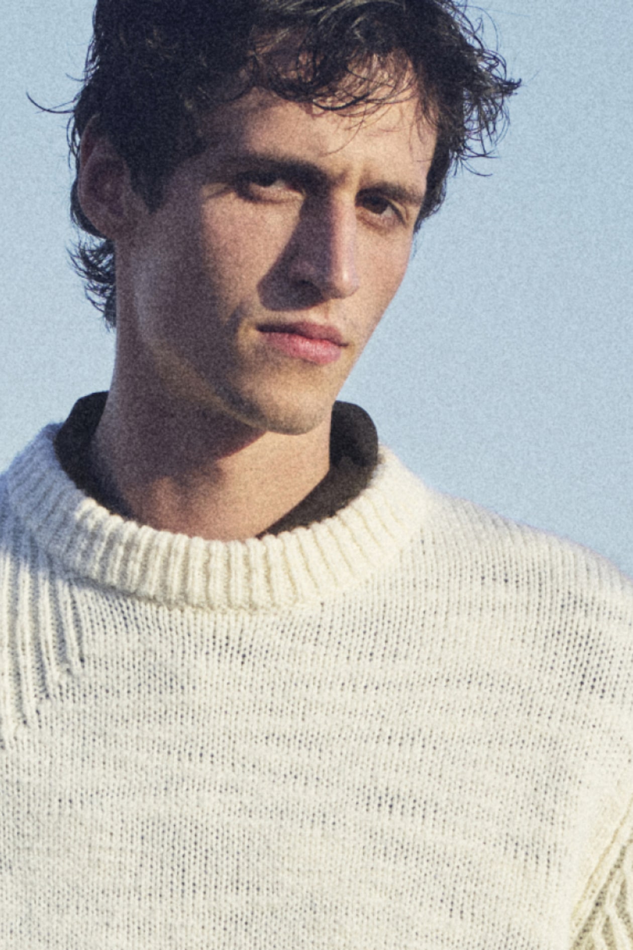 Person wearing a cream knitted sweater standing against a clear blue sky, looking slightly to the side with a calm expression.