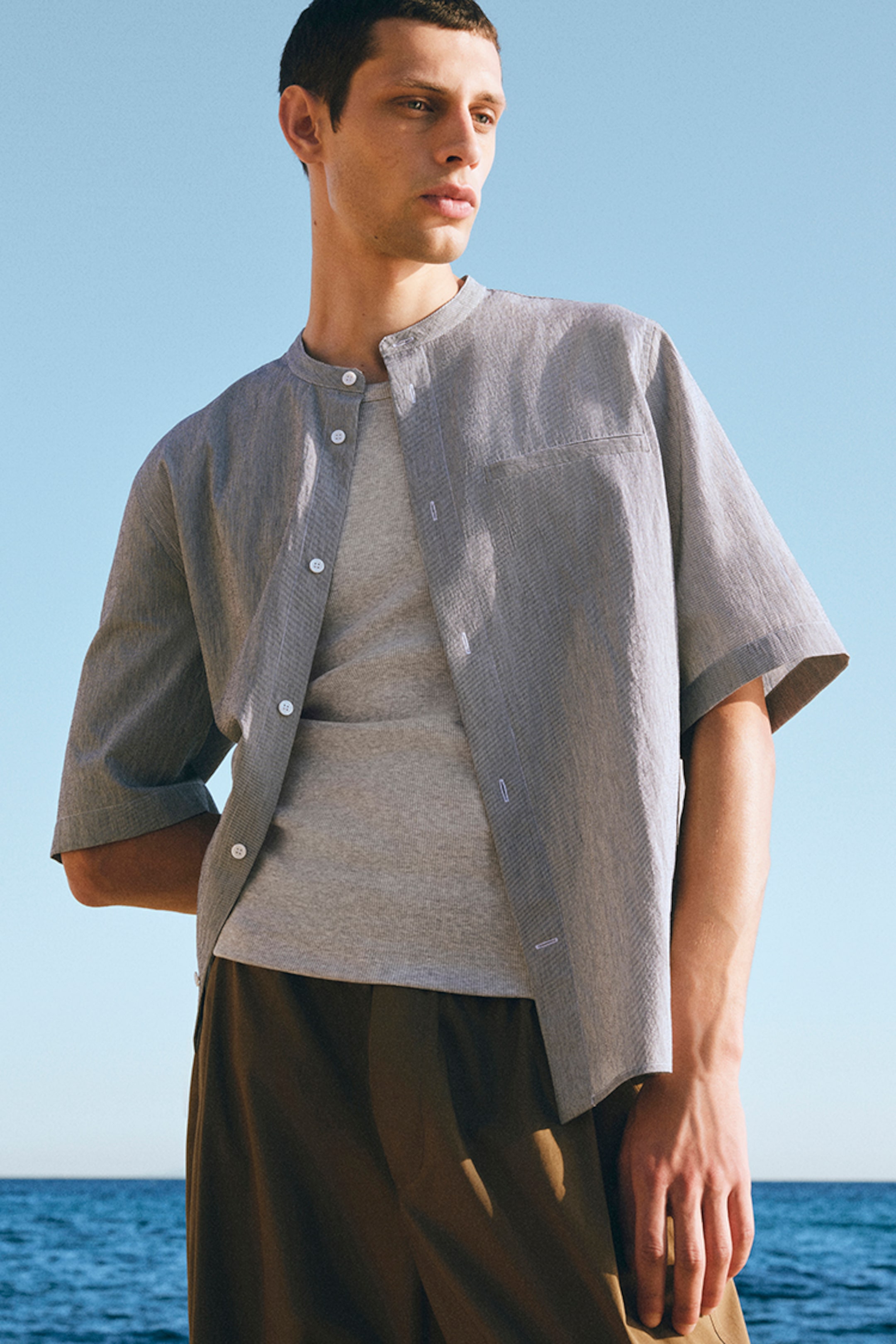 Man in a light gray shirt and brown pants stands by the sea, looking to the side under a clear blue sky.