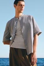 Placeholder for: Man in a light gray shirt and brown pants stands by the sea, looking to the side under a clear blue sky.