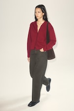 Placeholder for: A person wearing a red cardigan, dark pants, and black shoes walks while holding a brown bag. The background is a plain light color.