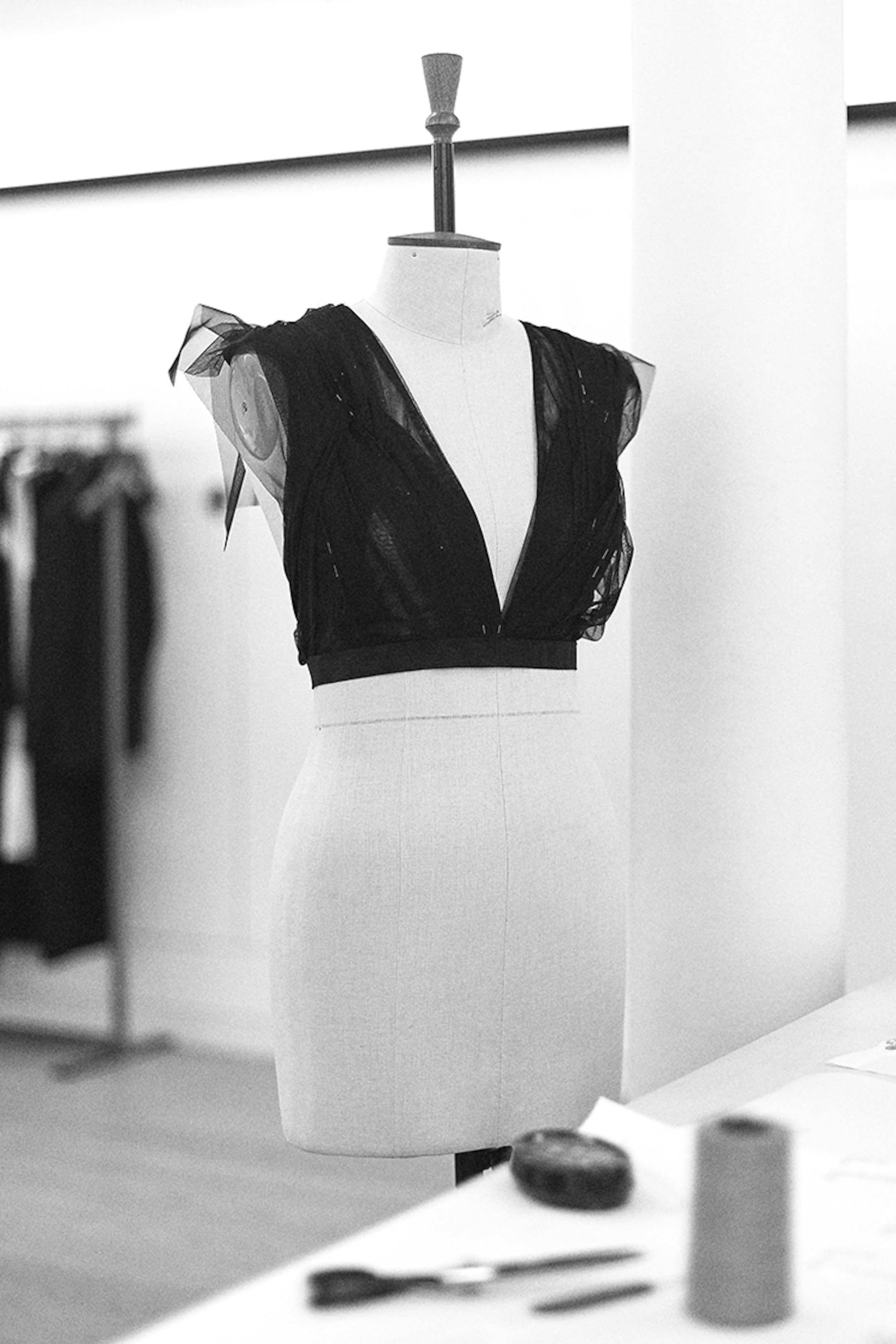 A dress form displays a black, sheer, sleeveless garment in a fashion studio. Scissors and thread are visible on a nearby table.