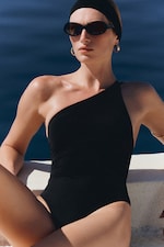 Placeholder for: Person in a black one-shoulder swimsuit, wearing sunglasses and hoop earrings, seated by the water with a serene expression.