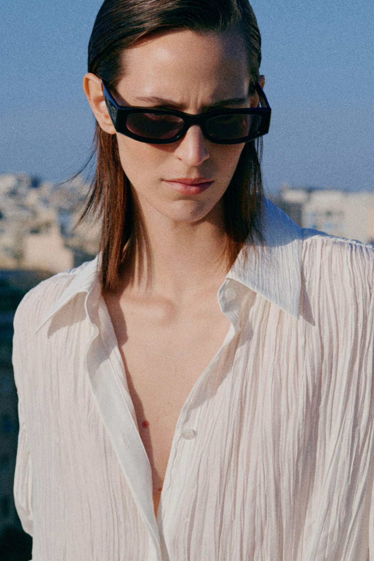 Person wearing sunglasses and a textured white shirt stands outdoors against a blurred cityscape under a clear blue sky.