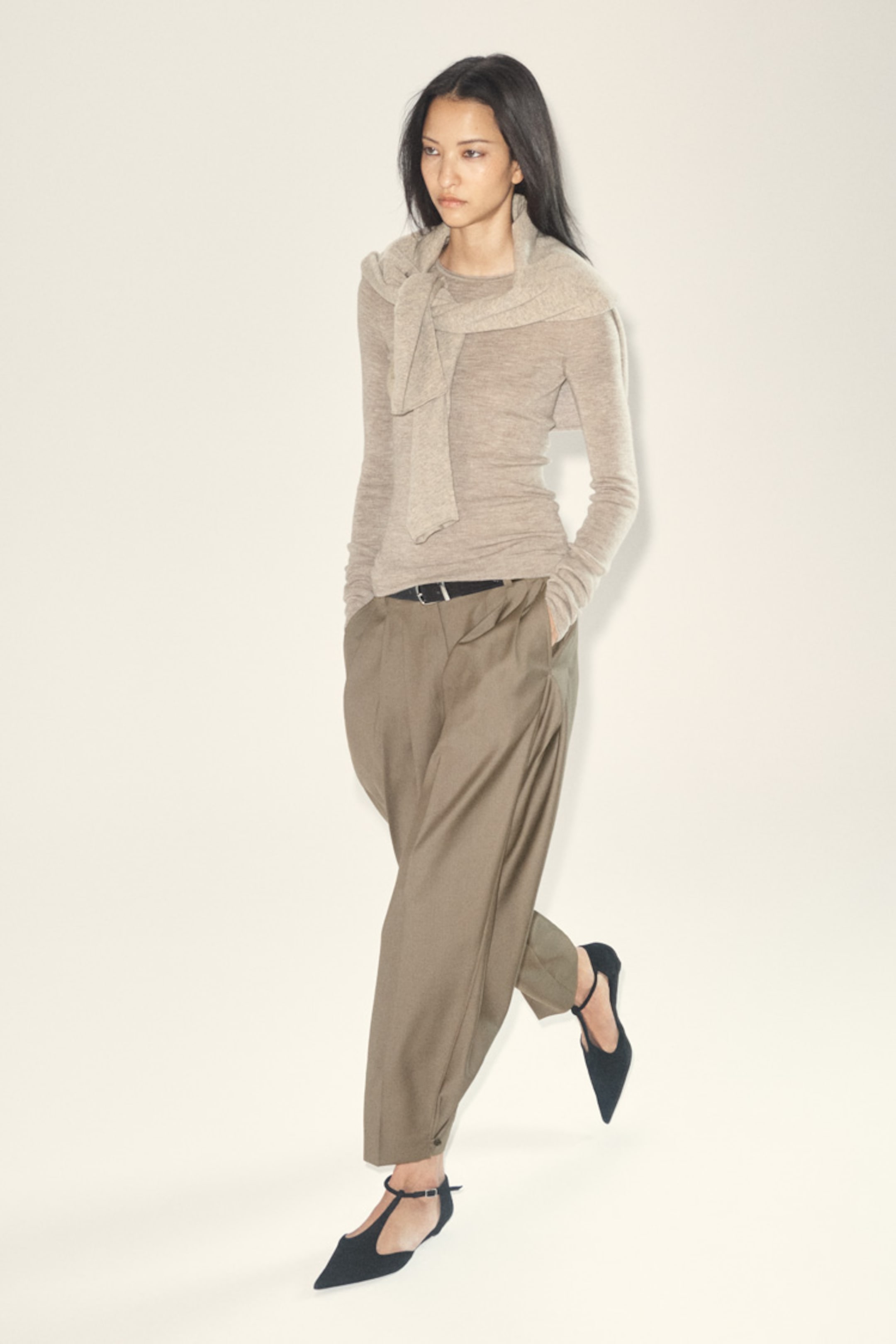 A person wearing a beige top and light brown pleated pants walks confidently. They have long hair and wear black pointed shoes.