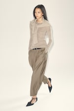 Placeholder for: A person wearing a beige top and light brown pleated pants walks confidently. They have long hair and wear black pointed shoes.