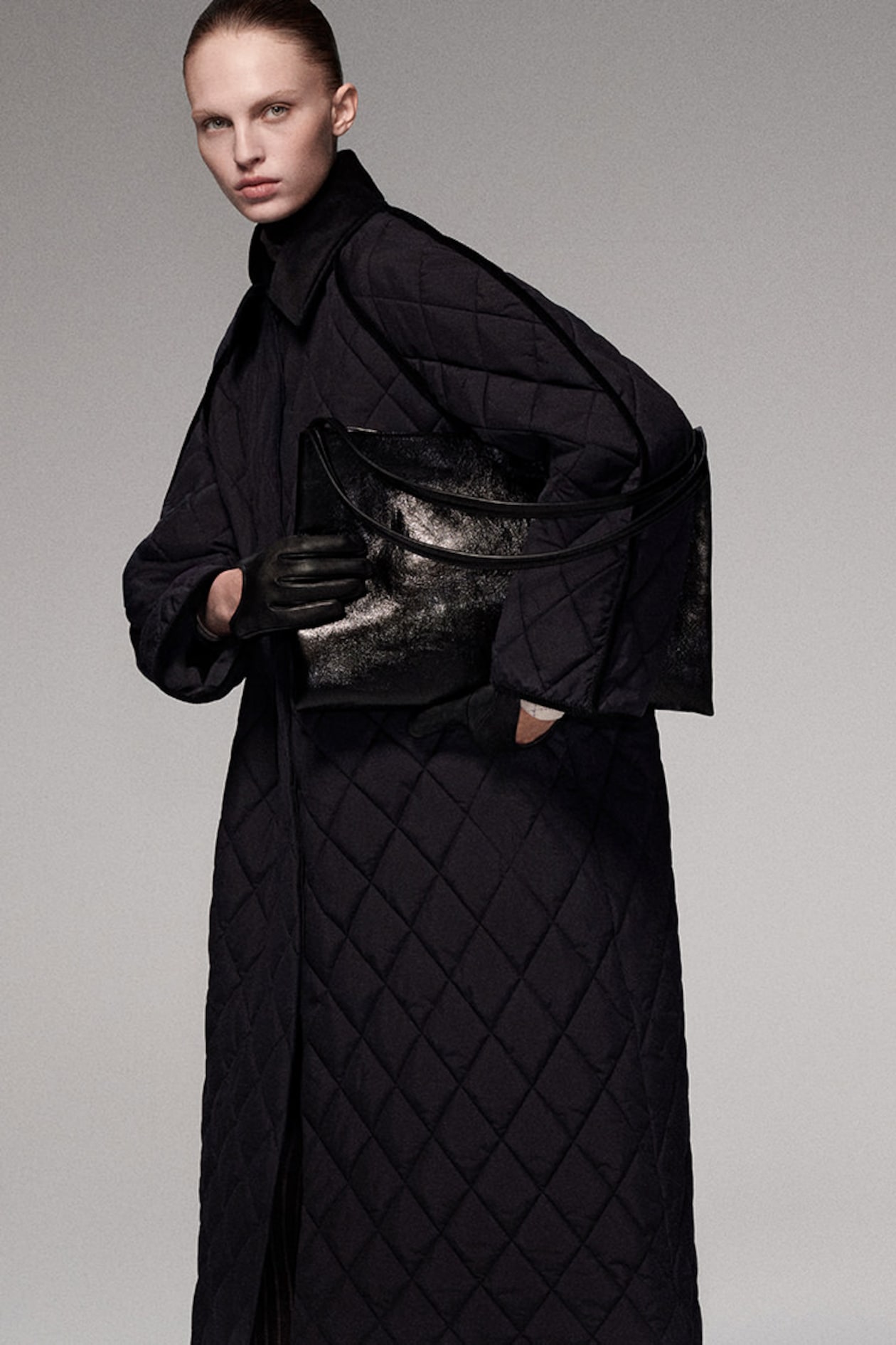 Person wearing a quilted black coat, holding a large black bag and wearing black gloves, standing against a neutral background.