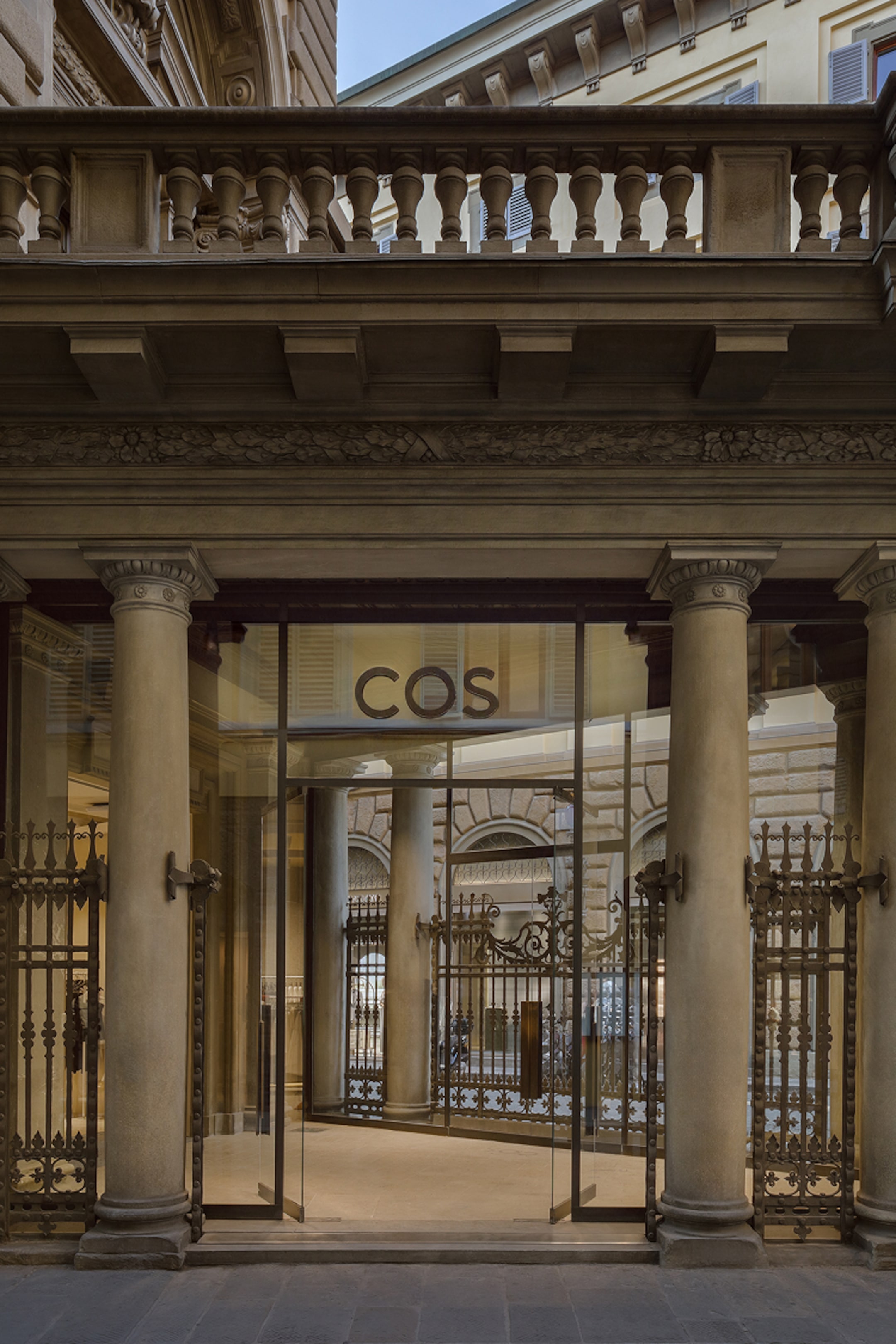 Entrance of a COS store with glass doors and ornate metal railings, set in a historic building with columns and detailed architecture.