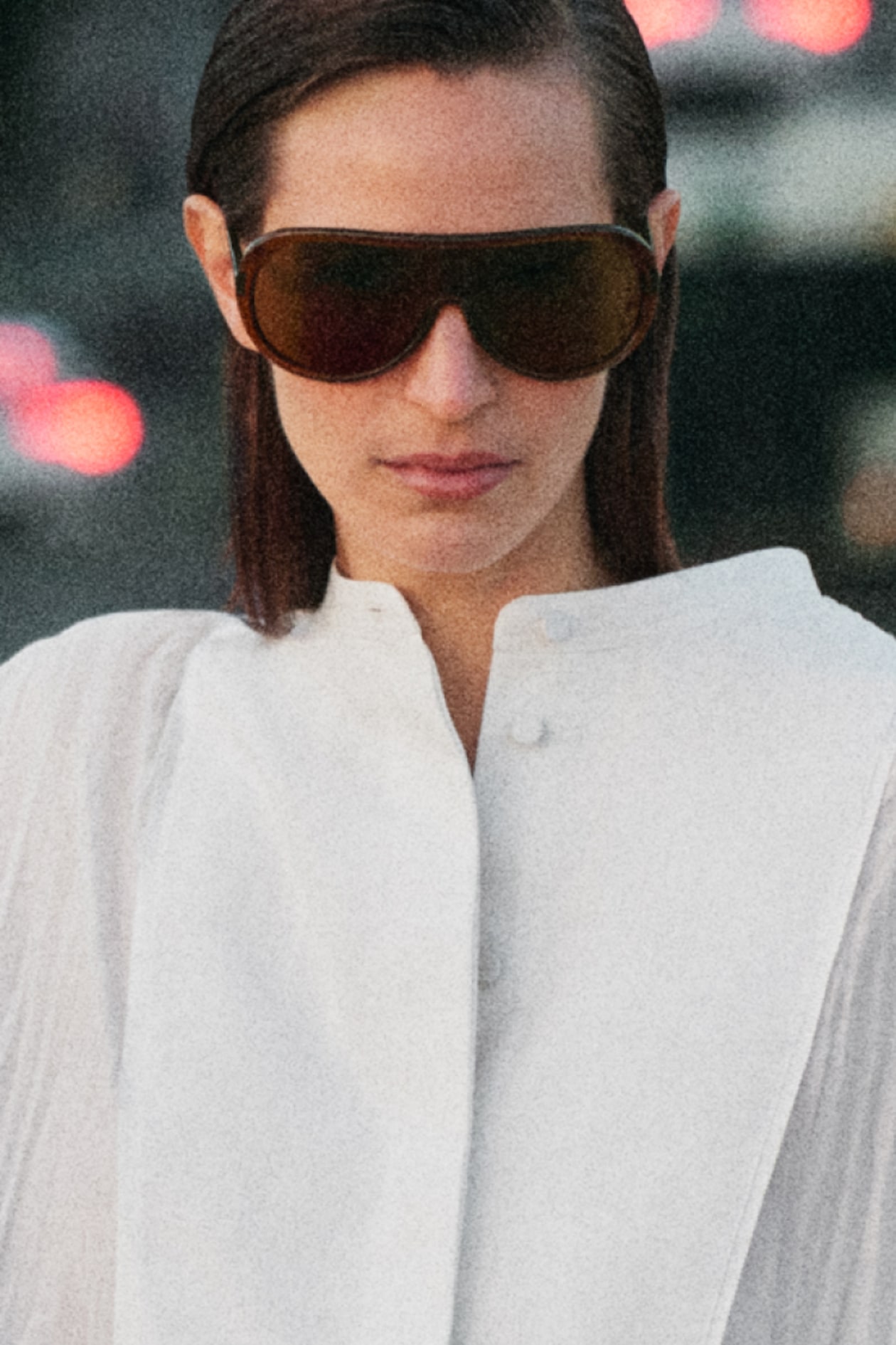 Person wearing oversized sunglasses and a white high-collared shirt, with blurred city lights in the background.
