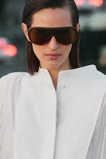Placeholder for: Person wearing oversized sunglasses and a white high-collared shirt, with blurred city lights in the background.