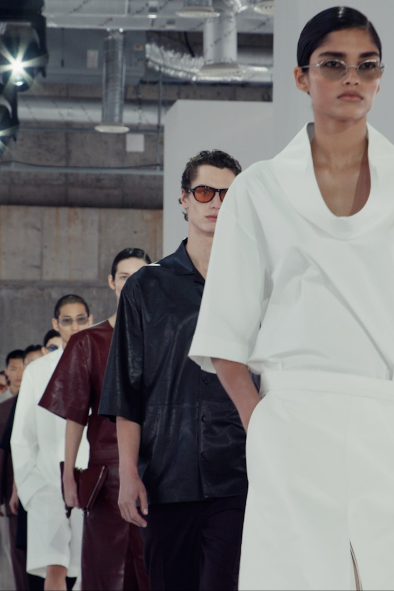 Models in minimalist outfits and sunglasses walking down a fashion runway, with industrial ceiling pipes visible above.