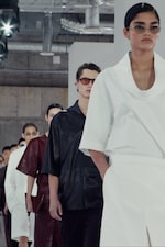 Placeholder for: Models in minimalist outfits and sunglasses walking down a fashion runway, with industrial ceiling pipes visible above.