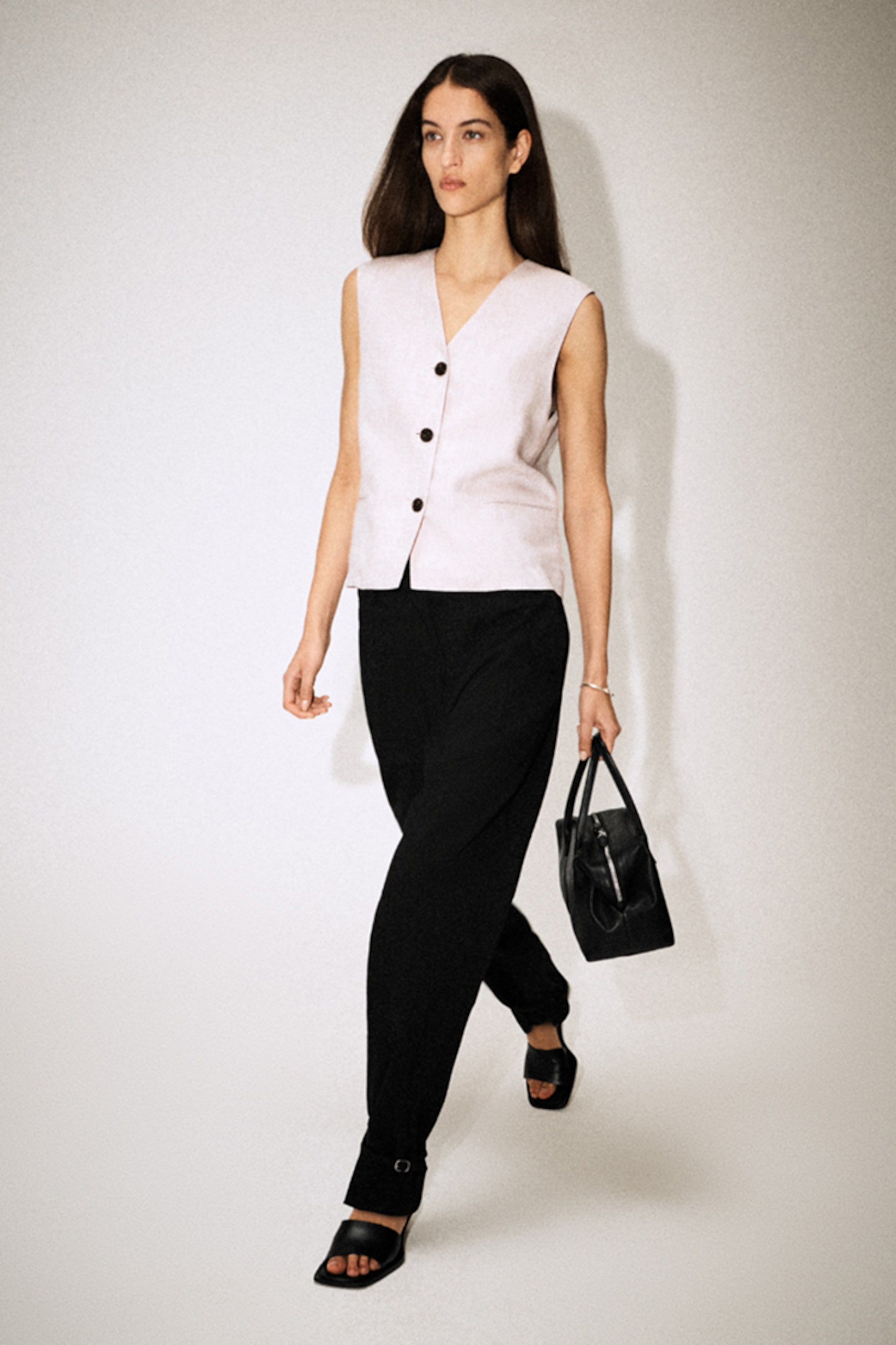 A woman with long hair wearing a sleeveless beige top and black trousers, carrying a black handbag, walks confidently on a light background.