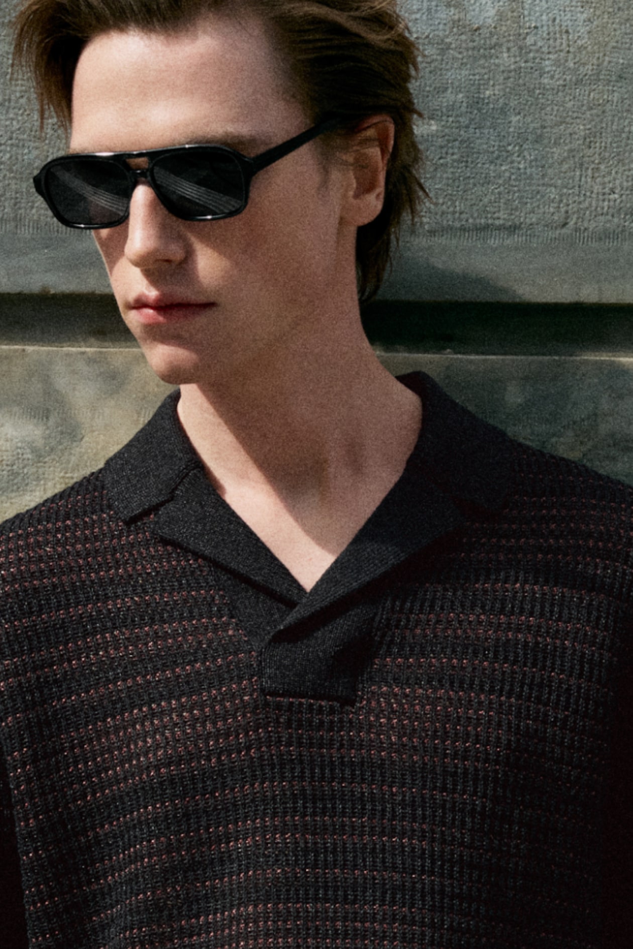 Person wearing black sunglasses and a textured knit shirt, standing against a stone wall, gazing to the side.