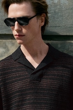 Placeholder for: Person wearing black sunglasses and a textured knit shirt, standing against a stone wall, gazing to the side.