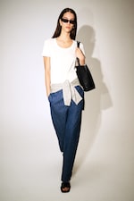Placeholder for: Woman in a white T-shirt, blue pants, sunglasses, and carrying a black bag, with a gray sweater tied around her waist.