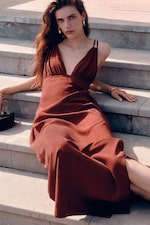 Placeholder for: Woman in a flowing rust-colored dress sits gracefully on stone steps, with long hair draped over one shoulder.