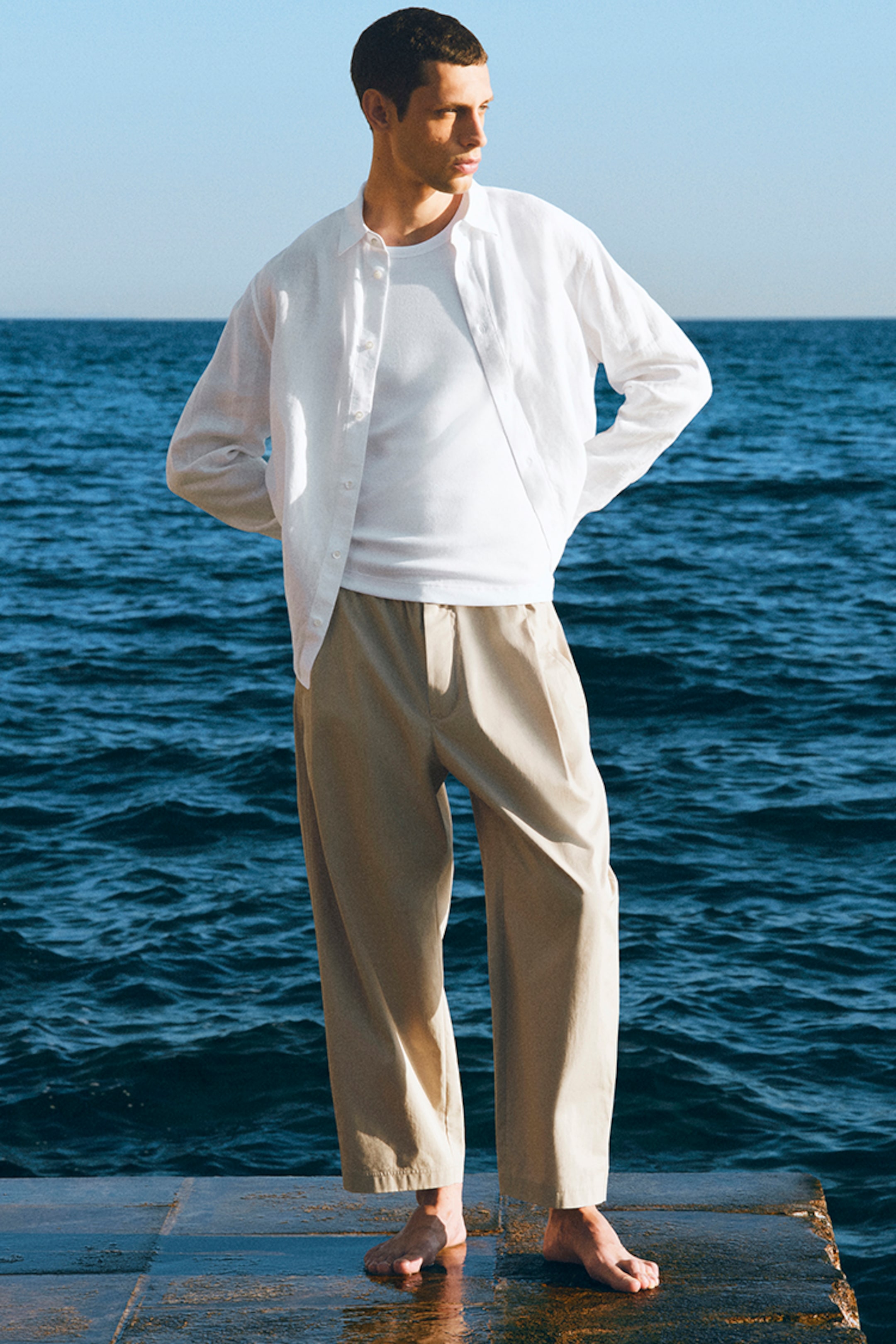 Man in a white shirt and beige pants stands barefoot on a pier, overlooking the sea under a clear blue sky.