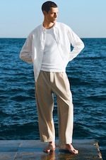 Placeholder for: Man in a white shirt and beige pants stands barefoot on a pier, overlooking the sea under a clear blue sky.