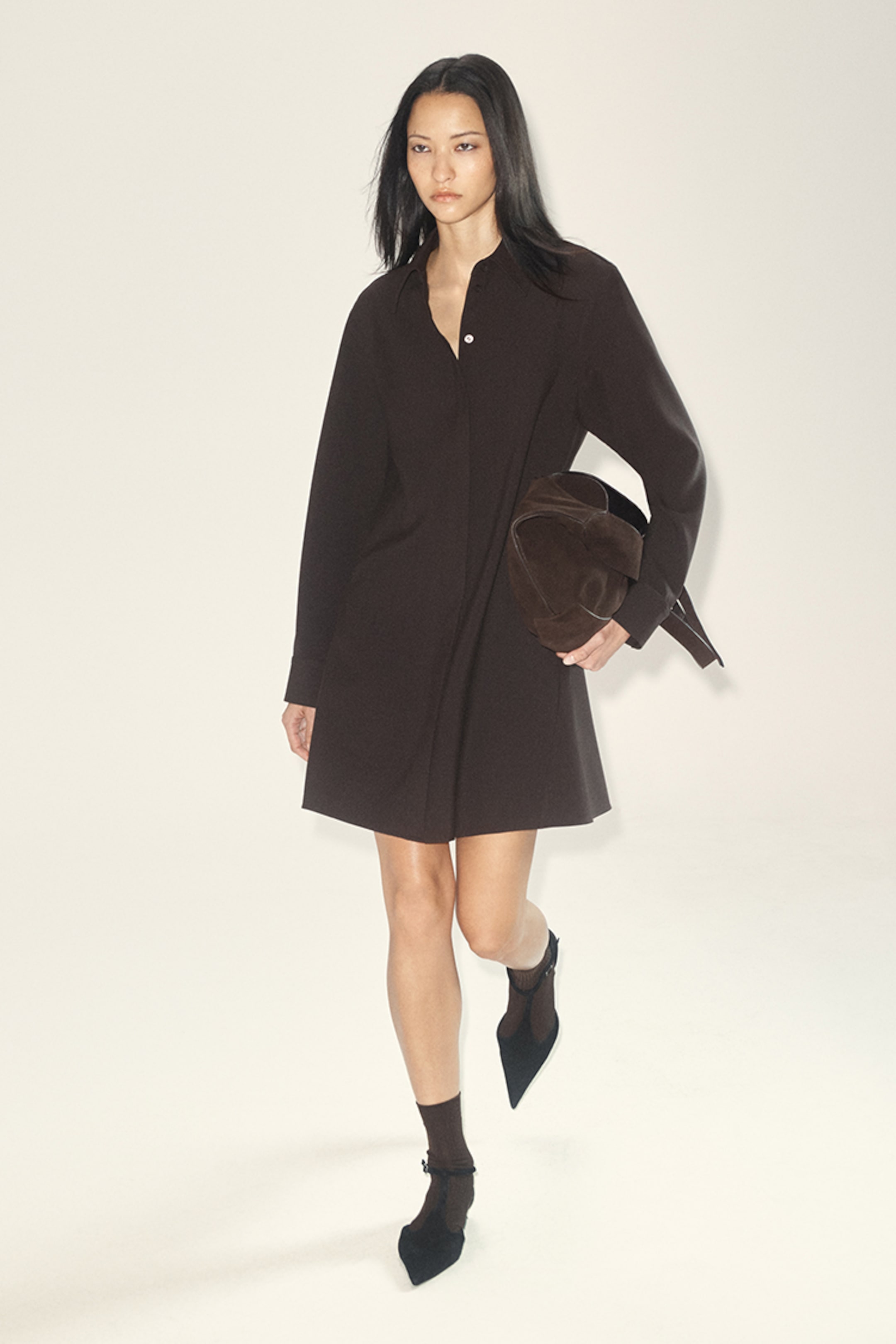 A woman in a dark long-sleeve dress and ankle boots walks, holding a matching hat, against a light background.